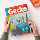 Magazin mit rotem Titelbalken und großer weißer Aufschrift „Gecko – Die Bilderbuchzeitschrift“ sowie der Ausgabennummer „107“. Darunter eine farbenfrohe Illustration mit anthropomorphen Stiften, einer Schere, einem Buch und weiteren Malutensilien mit Gesichtern vor hellblauem Hintergrund. Linke Hand und rechte Hand einer Person in grünem Strickpullover halten das Heft über hellem Tisch, rechts oben orangefarbene Ablageschale sichtbar.