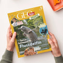 Zeitschrift „GEOlino mini“ mit gelbem Rand und Titelmotiv eines aus dem Wasser springenden Flussdelfins vor grünem Ufer. Textaufschriften: „Spitze Schnauze! Flussdelfin“, „KINDER Carissa bei den Bienen“, „WISSEN Komm mit auf Schatzsuche“ und „SUCHEN Alles in toller Unordnung“. Eine erwachsene Person hält das Heft mit beiden Händen über einer hellen Oberfläche, im Hintergrund eine orange Ablageschale.