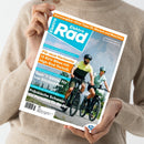Titelbild der Zeitschrift „ElektroRad“, gehalten von einer Person in beigefarbenem Pullover. Das Cover zeigt zwei radfahrende Personen in Sportkleidung und Helmen auf E-Bikes vor Berglandschaft. Sichtbare Texte: „ElektroRad“, „Für neue Horizonte – 13 SUV-Bikes mit High-End-Technik“, „Test: 11 Sättel für top Sitzkomfort“, „Fränkische Schweiz: Ein Paradies für Radler“, „Sommerglück: Hitze-Tipps für Mensch und Maschine“.