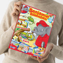 Eine hellhäutige Person in beigefarbenem Strickpullover hält eine bunte Kinderzeitschrift mit rotem Rand und dem Titel „Benjamin Blümchen“. Auf dem Cover ist der gezeichnete Elefant Benjamin in roter Kleidung und Mütze zu sehen, daneben bunte Illustrationen von Chamäleons, Gemüse-Snacks und einer Rechenaufgabe. Sichtbare Textabschnitte lauten u. a. „Gemüse-Snacks“, „So sehen Tiere“, „Doras Rechenpyramide“ und „Ein fliegendes Chamäleon“.