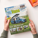 Titelseite des Magazins „Camper Vans“ mit hellblau-grünem Schriftzug. Darauf ein Foto eines blauen Campervans mit geöffnetem Aufstelldach, davor steht eine männliche Person mit grauem Haar in Jeans und hellem Oberteil. Im Hintergrund eine Wiese mit gelben Blumen. Sichtbare Schlagzeilen: „Der Multivan“, „Forster für fünf“, „Sprinter ohne 4x4“, „Mehr geht nicht“, „Tour de France – Mit dem Camper zur Tour“. Zwei Hände halten das Magazin über hellem Untergrund.