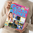 Person in beigefarbenem Strickpullover hält ein Magazin mit dem Titel „BASTEI GLÜCKSRÄTSEL“ mit pinkem Hintergrund und blauer Schrift. Auf dem Cover sind eine lächelnde Frau mit langen blonden Haaren, ein Rätselgitter und Abbildungen von Preisen wie Frühstücks-Sets, Tierpflege-Sets, Olivenöl-Sets, Freizeitpark-Tickets, Gläser-Sets und ein Urlaubsfoto zu sehen, dazu Textfelder in Gelb und Blau mit Hinweisen auf Gewinnaktionen.
