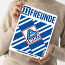 Weibliche Person im beigefarbenen Strickpullover hält ein schräg ausgerichtetes Magazin mit weißem Rand und blau-weißen Streifen. Auf dem Cover stehen „TIERFREUNDE“, „Magazin für Fußballkultur“, „BULLI“ und „SONDERHEFT 2026/27“; mittig befindet sich ein rot umrandetes BULLI-Logo.
