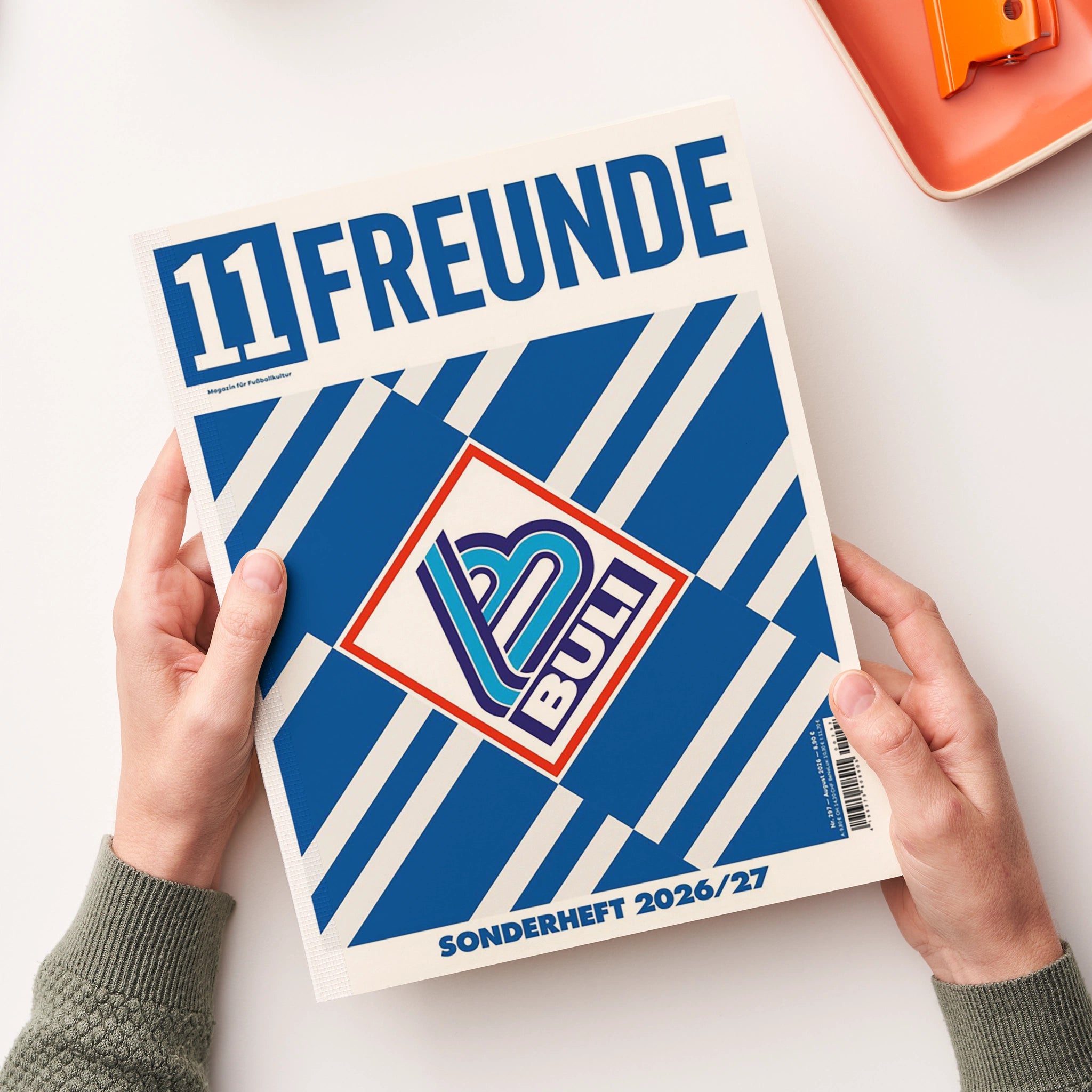 Blau-weiß gestaltetes Magazin „11 FREUNDE“ mit geometrischen Streifen und zentralem, mehrfarbigem Logo, von zwei Händen über einer weißen Oberfläche gehalten. Oben rechts liegt teilweise ein orangefarbenes rechteckiges Objekt mit abgerundeten Ecken.