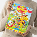 Person in beigefarbenem Strickpullover hält ein buntes Kinderheft mit dem Titel „Disney Winnie Puuh“. Auf dem Cover sind die Figuren Winnie Puuh, Tigger, Rabbit und Ferkel auf einer grünen Wiese zu sehen, umgeben von Bäumen und Vögeln. Unten befinden sich runde Bildfelder mit Motiven wie „Poster und Rätsel“ und „Naturforscher“, am unteren Rand steht in Rot und Gelb „Entdecken, Malen, Spielen“.