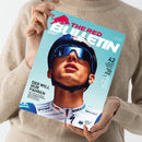 Person in beigem Strickpullover hält ein Magazin mit hellblauem Cover, Titel „THE RED BULLETIN“ und rotem Stier-Logo oben links. Abgebildet ist ein männlicher Radsportler mit Helm, verspiegelter Sonnenbrille und weißem Trikot mit Logos „BORA“, „grenke“ und „UCI WORLD TEAM“. Text auf dem Cover: „DER WILL NUR FAHREN“ und „42 SEITEN SOMMER-SPECIAL FEEL THE REAL“.
