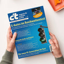Titelseite des Magazins „c’t magazin für computer technik“, Ausgabe 11, mit blauem Hintergrund. Haupttitel: „E-Autos im Reichweitentest“; Untertitel über Tests und Optimierung der Reichweite. Weitere sichtbare Rubriken: „Bluetooth-Audio nachrüsten“, „Die neue KI-Generation im Test“. Rechts eine Ampel mit grünem Licht und Blitzsymbol. Gehalten von zwei Händen über hellem Tisch mit orangener Schale oben rechts.