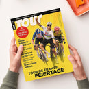 Titelblatt des Rennrad-Magazins „Tour“ in Gelbton mit drei männlich wirkenden Radrennfahrern in Rennkleidung und Helmen im Vordergrund, mittlerer Fahrer hebt den rechten Arm. Text auf dem Cover: „Tour de France Feiertage“, „64 Seiten! Extra-Heft zur Tour de France“, „Günstige Radhosen für Männer & Frauen“, „Auf den Spuren der Tour-Profis“, „Team Telekom“, „Die besten Touren am Arlberg“. Hände halten das Heft vor hellem Hintergrund.