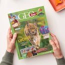 Titelseite des Magazins „GEOlino“ mit grünem Rand und großem Foto eines Luchses in natürlicher Umgebung. Oben links ein Foto von „Checker Tobi“ mit Schriftzug „NEU“ und „WERBUNG“, unten ein rechteckiges Foto von drei lachenden Jugendlichen. Weitere Texte: „Luchse – Zurück in unseren Wäldern“, „Vulkane – Wenn die Erde Feuer spuckt“ und „Auf der Balz“. Das Magazin wird von UNICEF unterstützt. Hände halten das Heft über hellem Tisch.