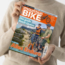 Eine Person in beigem Strickpullover hält das Magazin „mountainBIKE“ in den Händen; das Cover zeigt zwei männliche Radfahrer mit Helmen, Enduro-Mountainbikes und Berglandschaft im Hintergrund. Aufdrucke: „Das große Bosch-Update“, „Alles Enduro“, sowie Themen zu E-MTBs, Rucksack-Check und Touren. Farbgestaltung in Orange, Weiß und Blau, Magazinformat im Hochformat mit glänzender Oberfläche.