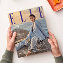 Titelblatt der Zeitschrift „ELLE“, Ausgabe Juni 2025, gehalten von zwei Händen über einer hellen Tischfläche. Auf dem Cover eine weiblich gelesene Person in hellblauem Blazer und Jeans vor Meereskulisse bei Sonnenuntergang. Sichtbare Texte: „GENERATION LOVE“, „BEAUTY MASTERCLASS“, „SOMMER DENIM“, „JETZT IM ELLE: STYLEGUIDEN, PERFEKTE SCHNITTE, NEUE SILHOUETTEN, PERFEKTE KOMBIS, WIR LIEBEN JEANS!“.