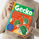 Person in beigem Strickpullover hält die Bilderbuchzeitschrift „Gecko“ (Nr. 114) mit grün-gelbem Titelbalken. Auf dem orangefarbenen Cover sind drei lächelnde Brokkoli-Figuren in weißen Raumanzügen schwebend im All dargestellt, begleitet von kleinen Raketen. Sichtbare Texte: „PIZZA sechs Jahreszeiten“, „JUPP und die Gemüsegäng“, „BROKKOLI im All“, „Geschichten, Sprachspiele und Mitmachseiten“, „Alle zwei Monate neu ohne Werbung“, „THEMA Gemüse!“.
