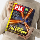 Person in beigefarbenem Strickpullover hält ein Exemplar des Magazins „P.M.“ mit gelbem Rand und rotem Logo. Das Titelthema lautet „Neue Super-Teleskope“ in großen gelben Buchstaben, gezeigt wird ein großes Observatorium mit mehreren orange leuchtenden Laserstrahlen am Nachthimmel. Weitere sichtbare Schlagzeilen: „Bahn & KI“, „Hinter Gittern“, „Kaufe Dino“.