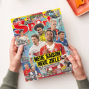 Titelbild der Zeitschrift „BRAVO Sport“ mit gelbem Hintergrund; mittig mehrere männliche Fußballspieler in verschiedenen Vereins-Trikots, darunter rot-weiß, gelb-schwarz und blau, in Halbporträts angeordnet. Große Überschrift „NEUE SAISON NEUE ZIELE“ in Schwarz-Gelb, weitere Textelemente wie „KRASSE EXTRAS!“, „HEFTIGE BMX-ACTION“ und „SAISONSTART IN LIGA 2“. Gehalten von zwei Händen vor hellem Tischhintergrund.