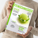 Helle Zeitschrift „Feinschmecker“ mit grün-weißer Titelseite, gehalten von einer Person in beigefarbenem Strickpullover; mittig abgebildet ist eine runde, hellgrüne Dessertkugel auf weißem Teller, umgeben von Kräutern. Sichtbare Textausschnitte: „Jetzt an die Côte d’Azur!“, „Sommerküche“, „Grüne Welle – 30 Seiten über Olivenöl“, „Hamburg – Koch des Monats: Maurizio Oster“.