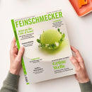 Titelblatt der Zeitschrift „Feinschmecker“ mit hellgrün-weißer Gestaltung, mittig ein kugelförmiges grünes Dessert auf weißem Teller, dekoriert mit Kräutern und Blüten. Textaufschriften: „Jetzt an die Côte d’Azur!“, „Sommerküche“, „Gardasee“, „Kaffee-Metropolen“, „Hamburg“, „Grüne Welle“. Gehalten von zwei Händen über weißer Tischfläche, im Hintergrund orangefarbene Schale mit Messer.