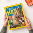Magazin „National Geographic Kids“ mit gelbem Rand und blauem Titelhintergrund, zentral ein Leopardenfoto mit der Überschrift „DSCHUNGEL: Leoparden im Regenwald!“. Linke Spalte mit kleineren Bildern zu Themen wie Gepard, Papageien, Lemuren und Strandtieren. Unten rechts ein gelber Hinweis „2 tolle Poster!“. Gehalten von zwei Händen über hellem Tisch mit orangefarbenem Objekt oben rechts.