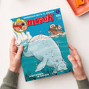 Comic-Heft mit der Titelaufschrift „mosaik – Unterwegs mit G. W. Steller“, Ausgabe 594, Juni 2025. Das farbige Cover zeigt eine gezeichnete graue Seekuh unter Wasser vor Eisbergen, darüber ein Boot mit einer in Pelz gekleideten Person. Im unteren Bereich steht der Text „Schiffbruch im Eismeer“. Zwei Hände in grünem Strickpullover halten das Heft über einer hellen Oberfläche mit orangefarbener Ablage.