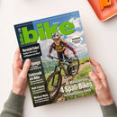 Titelblatt der Zeitschrift „bike“ Ausgabe 06/2023 mit großem grünem Schriftzug „bike“ oben. Im Mittelpunkt ein weiblich gelesene Person mit Helm, Sonnenbrille und Radbekleidung, die auf einem Mountainbike bergauf fährt, umgeben von grüner Landschaft und blauem Himmel. Sichtbare Schlagzeilen: „Vorsicht Fake!“, „Elektronik am Bike“, „4 Spaß-Bikes im Vielseitigkeits-Check“, „Dolomiten – Die besten Touren im Eggental“. Hände halten das Magazin über hellem Tisch.