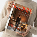 Person in beigem Strickpullover hält ein Magazin mit dem Titel „GOODLIFE WOHN-DESIGN“. Auf dem Cover steht eine männlich gelesene Person in dunkler Kleidung in einem modern eingerichteten Raum mit Holztisch, Stuhl, Pflanze und Dekogegenständen vor einer braunen Wand. Sichtbare Texte: „INTERIEUR“, „KULTUR“, „GENUSS“, „REISEN“ sowie „N°187 RÄUME MIT TIEFGANG – Entdecken Sie mit uns neue Einrichtungsstile, Materialien und kreative Gestaltungsansätze“.