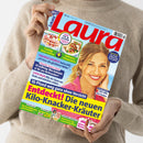 Titelblatt der Zeitschrift „Laura“ mit großem roten Schriftzug auf blauem Hintergrund; rechts im Vordergrund eine lächelnde Frau mit blondem Haar in rosa Hemd. Um sie herum mehrere farbige Textfelder mit Schlagzeilen zu Themen wie Ernährung, Gesundheit, Einkaufen und Sommermode. Oben Bilder von zwei Gerichten mit den Beschriftungen „Fruchtige Baiserkuchen“ und „Raffinierte Urlaubsküche“. Gehalten von einer Person in beigefarbenem Strickpullover.