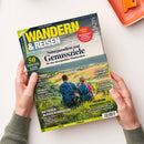 Titelbild des Magazins „Wandern & Reisen – Sommer 03-26“. Zwei Personen mit Rucksäcken sitzen auf einer grünen Wiese mit Blick auf ein Tal und ein Dorf. Hauptüberschrift: „Naturparadiese und Genussziele für eine unvergessliche Wandersaison“. Weitere Texte: „50 frische Ideen“, „Ratgeber: Blasen verhindern“, „Premium-Wanderwelten“, „Osterspaier Langhalsweg“ und „Deutschland lockt“. Hände halten das Heft über hellem Untergrund.