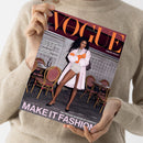 Magazincover der deutschen Ausgabe „VOGUE“ mit orangefarbenem Schriftzug und Texten wie „LANGES HAAR RICHTIG PFLEGEN“, „WEDDING-SPECIAL: MODERNE HOCHZEITSLOOKS“ und „MAKE IT FASHION“. Auf dem Titel steht eine weibliche Person mit langen dunklen Haaren, hellrosa Mantel, orangen Handschuhen und hellen Shorts vor einem Hintergrund mit Bistrostühlen und Marmormauer. Gehalten von einer Person in beigefarbenem Pullover.