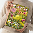 Person in beigem Strickpullover hält das Magazin „kraut&rüben – Schöner. Wilder. Biogarten.“ (Ausgabe 07/2026) vor neutralem Hintergrund; Titelbild zeigt eine Nahaufnahme von gelben und pinken Blüten auf einer Sommerwiese. Sichtbare Texte: „TOMATEN-LIEBE – Pflege-Leitfaden für köstliche Früchte“, „GRIECHISCHES SEIN“, „ZUKUNFTSSPEICHER“, sowie Hauptüberschrift „Jeder Tag ein Sommerfest!“.
