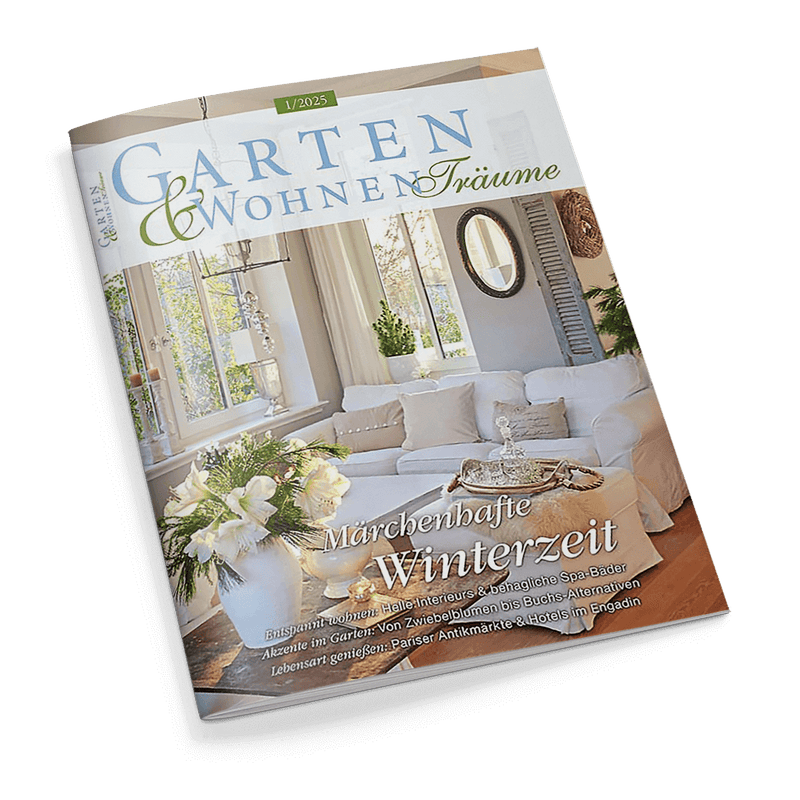 Titelbild der Zeitschrift „Garten & Wohnen Träume“, Ausgabe 1/2025, mit einem hell eingerichteten Wohnzimmer im Landhausstil: weiße Sofas, große Fenster, Spiegel, Holzelemente und Blumenarrangement in weißer Vase im Vordergrund; sichtbare Texte am unteren Rand u. a.: „Märchenhafte Winterzeit“, „Entspannt wohnen“, „Akzente im Garten“ und „Lebensart genießen“.