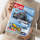 Person in beigefarbenem Pullover hält die Juli-2026-Ausgabe des Luft- und Raumfahrt-Magazins „FLUG REVUE“, Titelstory „Fliegende Panzer – Ungarns letzte Mi-24“. Auf dem Cover ein grauer Kampfhubschrauber vor blauem Himmel, darunter kleinere Bilder mit einem gelben Passagierflugzeug beim Start und einem Raumfahrtsmotiv; sichtbare Schlagzeilen zu Militärtechnik, Luftfahrtkrise und Energie im All.