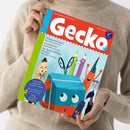 Person in beigem Strickpullover hält die Kinderzeitschrift „Gecko“ (Ausgabe Nr. 107) mit rotem Titelkopf und weißem Schriftzug. Das Cover zeigt eine farbenfrohe Illustration mit einem blauen Buch mit Augen, einer Schere, einem Bleistift und bunten Stiften mit Gesichtern. Aufgedruckte Texte: „Die Burg Mackefei“, „Angestiftet“, „Der Tag des fröhlichen Wurms“ sowie ein blauer Kreis mit „Geschichten, Sprachspiele und Mitmachseiten“.