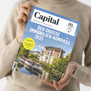 Person in beigefarbenem Strickpullover hält die Mai-Ausgabe 2023 des Wirtschaftsmagazins „Capital“ mit dem Titelthema „Der große Immobilien-Kompass 2025“. Auf dem Cover: ein Foto eines weißen Hauses mit rotem Dach am Wasser, davor ein Ausflugsboot, umgeben von Bäumen unter blauem Himmel. Sichtbare Textelemente u. a. „TRUMP-CRASH“, „Aktien, Anleihen, Gold“, „Vertiefung“ und „Erfolgs-Start-up“.