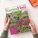 Magazintitel „GartenFlora“ Juni 2025 mit dem Untertitel „gärtnern, erleben und genießen“, gehalten von zwei Händen über hellem Hintergrund; Titelfoto zeigt einen üppigen Garten mit rosafarben blühenden Rosenbüschen, grünem Laub und einem Weg im Hintergrund; kleineres Bild oben links zeigt eine Topfpflanze mit Sukkulente; Textausschnitte u. a.: „Die Besten für den Garten – Duftrosen“, „Gartenkunst“, „Rasenmäher mit Akku-Antrieb“, „Rezepte mit Süßkirschen“.