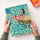 Zwei Hände halten eine Ausgabe des Magazins „flow“ mit blauem Hintergrund und dichtem Muster aus gelben und weißen Gänseblümchen; mittig großes rosa Schriftlogo „flow“, darunter eine Illustration einer weiblichen Figur in einem Boot voller Blumen mit der Aufschrift „Man muss das Unmögliche üben, um das Mögliche zu erreichen“, oben links Kreis mit Text „5 Lese-, Bastel- & Büchlein“.