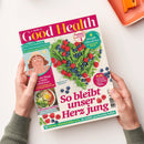 Person mit hellbeigem Hautton hält eine Ausgabe der Zeitschrift „Good Health“ vom Mai/Juni 2025, Titel in roter Schrift auf pinkem Hintergrund. Auf dem Cover sind ein herzförmig angerichteter Mix aus grünen Bohnen, Himbeeren und Blaubeeren sowie Texte wie „So bleibt unser Herz jung“, „Das Einmaleins des Sonnenschutzes“ und „Straffe Oberarme“ zu sehen, ergänzt durch kleinere Bildfelder mit Gesichtsillustration und Frühstücksschüssel.