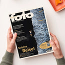 Titelblatt des „fotoMAGAZIN“ August 2026 mit der Aufschrift „Die ganze Welt der Fotografie“. Linke Seite mit schwarzem Hintergrund und weiß-gelber Schrift zu Themen wie „Drohnenfotografie“, „Michael Kenna“, „Leichtes Gepäck“ und „Schöne Reise!“. Rechts zeigt eine Luftaufnahme eine Person in dunkler Badehose am Rand brechender Meereswellen, gehalten von zwei Händen über hellem Tisch mit orangener Schale.