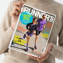 Person in beigem Strickpullover hält das Magazin „Runner’s World“ mit dem Titel in weißer Schrift auf grauem Hintergrund. Auf dem Cover laufen eine weibliche und eine männliche Person in violetten Sportoutfits und Sonnenbrillen nebeneinander. Sichtbare Textelemente: „Laufen Next Level“, „Das wird dein Sommer“, „Welcher ist der beste Laufschuh?“, „Endlich der richtige Sport-BH“, „Champion-Couple“ und weitere Themen rund ums Laufen.