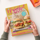 Person mit hellen Händen hält das Magazin „slowly veggie! Vegetarisch & vegan genießen“ mit gelbem Cover, pink-lila Logo und großem Titeltext „Burger, Wraps & Bowls“. Abgebildet sind zwei belegte Burger mit Sesambrötchen, Gemüse und Sauce auf hellem Teller. Weitere sichtbare Texte: „68 Gute-Laune Rezepte für den Sommer“, „Beeren-Saison“, „Coolcation“, „Smokey Jackfruit-Burger mit Coleslaw“. Hintergrund hell, Tischoberfläche beige.