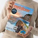 Person in beigem Strickpullover hält das Magazin „outdoor – Wandern. Reisen. Abenteuer.“ mit Titelbild einer hohen Felsküste bei Sonnenuntergang, einer Person auf dem Klippenrand und blauem Zelt im Vordergrund; sichtbare Schlagzeilen: „Die schönsten Küstentouren“, „Peaks of the Balkans“, „Wildes Trentino“, „Biber, Schilf & Seeadler“, „Zelte im Sturm“ und „Die besten Produkte 2026“.
