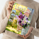 Person in beiger Strickjacke hält ein Magazin mit dem Titel „Landlust“. Das Cover zeigt einen Strauß aus violetten, rosa und weißen Blumen vor unscharfem grün-gelbem Hintergrund. Textaufschriften: „Sommer-Geschenke“, „Selbst gebaut: Pflanzkasten für Kräuter“, „Fruchtig: Kuchen ohne Backen“, „Rettich anbauen und ernten“. Linke Spalte mit kleineren Fotos von Landschaft, Haus, Garten und Schafen sowie grünem Schriftzug „Gewinnspiel“ oben links.