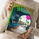 Person in beigem Strickpullover hält das Magazin „AW Architektur & Wohnen“ mit grün-goldenem Cover. Titelzeilen: „Unterwegs Mailand for Design-Fans“, „Dossier: Erlebt der Plattenbau einen Imagewandel?“ und „Das Beste aus Italien“. Abgebildet ist ein modern eingerichtetes Wohnzimmer mit blauem Sofa, runder Wandöffnung, Pflanzen und hellem Holzboden, aufgedruckter Kreis mit Text „AW Architekt des Jahres 2026“.