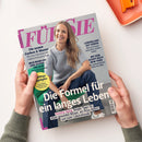 Titelblatt der Zeitschrift „FÜR SIE“ mit einer lächelnden weiblichen Person in grauem Pullover und blauer Hose vor grauem Hintergrund, sitzend auf einem grünen Möbelstück. Sichtbare Texte: „Die Formel für ein langes Leben“, „Healthy Aging: Simple Tipps für einen gesunden Körper, ein fittes Gehirn und mehr Lebensfreude“, Themen zu Farben, Mustern, Beziehungen, Reisen und Gesundheit. Zwei Hände halten das Magazin über einer hellen Oberfläche.