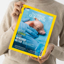 Person in beiger Strickpullover hält eine Ausgabe des Magazins „National Geographic“ mit gelbem Rand und blauer Titelseite. Auf dem Cover liegt ein kleines rosafarbenes Ferkel auf blauem Tuch, gehalten von Händen in blauen Latexhandschuhen. Hauptüberschrift: „Dieses Schwein kann Menschen retten“. Oben steht „Juni 2024“, weitere kleinere Texte zu Themen wie Baummeister, Zwergplanet, Inseln und Regenbogen.