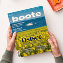 Titelblatt der Zeitschrift „boote“ Ausgabe 6/2025, gehalten von einer Person in olivgrünem Strickpullover über hellem Untergrund. Das Cover zeigt im oberen Bereich ein Motorboot auf blauem Meer, im Vordergrund ein gelbes Rapsfeld. Sichtbare Texte: „Gebrauchtboote“, „Wakeboard“, „Revier-Update Ostsee“, „Törnplanung“, „Führerschein“, sowie Länderhinweis „Deutschland, Polen und Dänemark“.