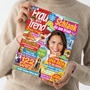 Helle Zeitschrift „Frau im Trend“ mit farbenfrohem Cover, im Vordergrund eine lächelnde Frau mit braunen Haaren vor türkisfarbenem Hintergrund. Schlagzeilen u. a.: „Schlank bis zum Urlaub“, „3 Hefte 1 Preis!“, „122 Pfund weg“, „Heiße Spar-Ideen für den Sommer“, dazu Bilder von Himbeertorte, Koteletts und weiteren Personen in kleineren Porträts. Gehalten von einer Person in beigefarbenem Strickpullover vor neutralem Hintergrund.