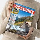 Person in beigem Pullover hält das Magazin „ROADBIKE“ mit rotem Titelkopf und blauem Hintergrund. Auf dem Cover fahren vier Radfahrer in bunter Rennbekleidung eine asphaltierte Bergstraße entlang, umgeben von grünen Hängen und blauem Himmel. Sichtbare Texte: „LEICHT & SCHNELL“, „FITNESS“, „Elektro-Pumpen“, „Sport-BHs“, „SCHÖNES ITALIEN“ sowie Themen zu Tests, Training und Radsport-Ausrüstung.