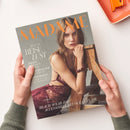 Titelblatt der Zeitschrift „MADAME“ in orangefarbener Schrift, mit einer weiblichen Person in weinroter Spitzenbekleidung, die mit dem Arm auf eine hölzerne Struktur gestützt sitzt; grauer Hintergrund, sichtbare Texte: „SPECIAL REISELUST“, „JAPANS SCHÖNSTE ORTE“, „GRIECHENLAND DER INSIDER“, „KOPENHAGEN, EUROPAS STILMETROPOLE“ und „BEACH-WEAR & ACCESSOIRES MIT OCEAN FLAIR“. Hände halten die Zeitschrift über heller Oberfläche.