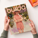 Titelblatt der Zeitschrift „burda style“ mit einer weiblich gelesenen Person in rosafarbenem Kleid, großem Strohhut mit Band und Sonnenbrille vor grünem Pflanzenhintergrund. Aufschriften u. a.: „La Dolce Vita“, „Sporty oder Couture“, „Exklusive Männerschnitte“, Hinweis auf 75 Jahre burda style und das Thema „Looks für einen unvergesslichen Sommer“. Zwei Hände halten das Magazin über hellem Tischhintergrund.