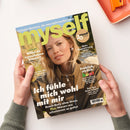 Titelbild der Zeitschrift „myself“ mit hellgrünem Logo und goldfarbenem Kreis „20 Jahre“. Im Mittelpunkt: eine blonde Frau in olivgrüner Jacke mit Reißverschluss, neutraler Gesichtsausdruck, vor unscharfem Naturhintergrund. Textaussagen u. a.: „Ich fühle mich wohl mit mir“, „Willst du irgendwann Kinder?“, „Fröhlich, stylish, bunt“ und „Mehr Begeisterung, bitte!“. Hände in olivgrünen Ärmeln halten das Magazin über hellem Tisch.
