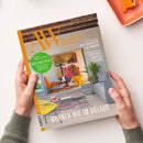 Titelbild der Zeitschrift „AW Architektur & Wohnen“ mit orangem Logo oben links, zentralem Foto eines modern eingerichteten Wohnraums mit buntem Wandbild, orangefarbenem Stuhl, gelbem Tisch, grauem Sofa, grüner Pflanze und farbig gemustertem Teppich. Textaufschriften u. a.: „52 Seiten Bad & Wellness“, „Neue Architektur aus Dänemark“, „Kunstvolle Muster & Texturen“, „Wohnen wie im Urlaub“. Zwei Hände halten das Magazin vor hellem Hintergrund.